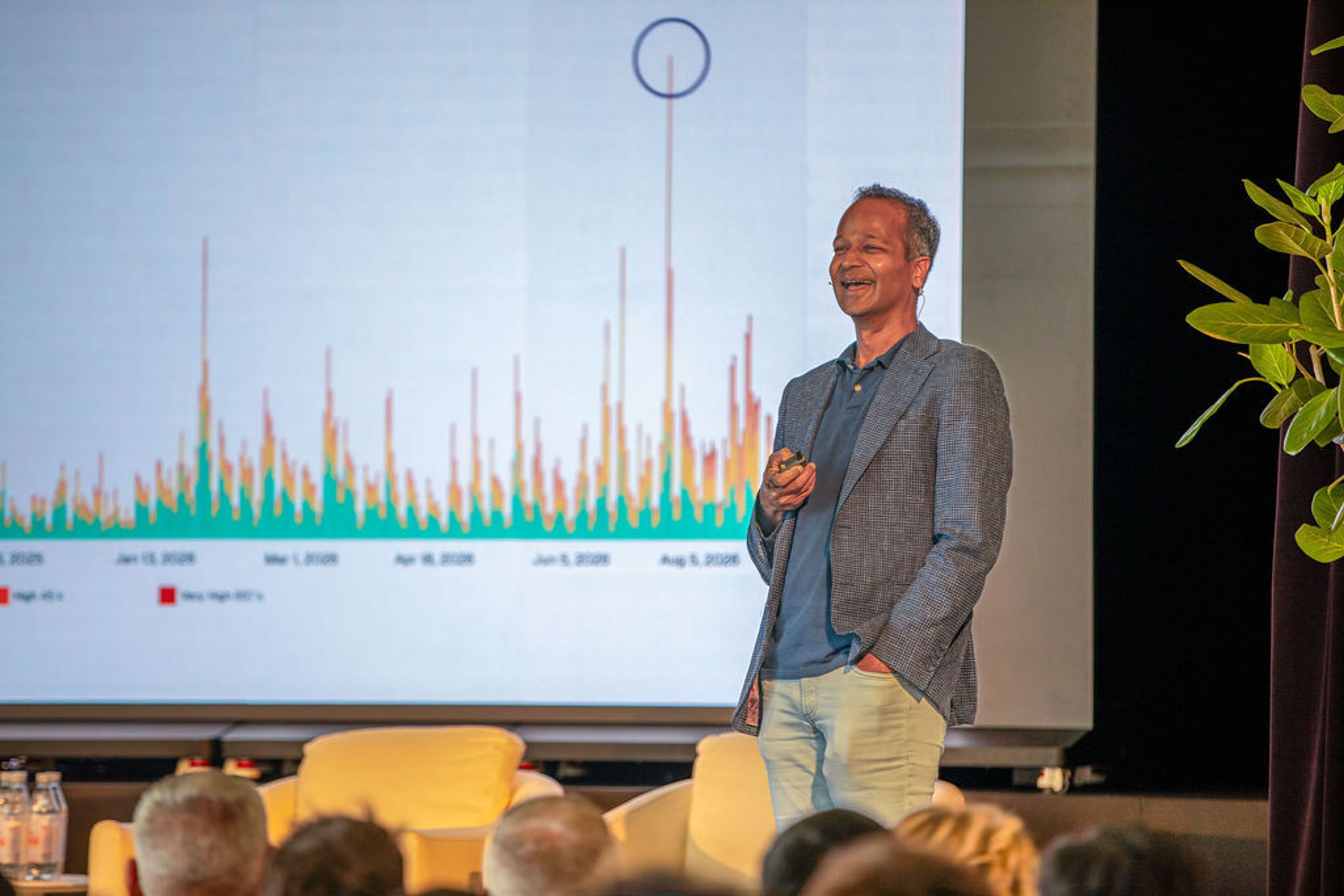 A smiling man presents on stage in front of a large screen displaying a bar chart, with an audience in the foreground.