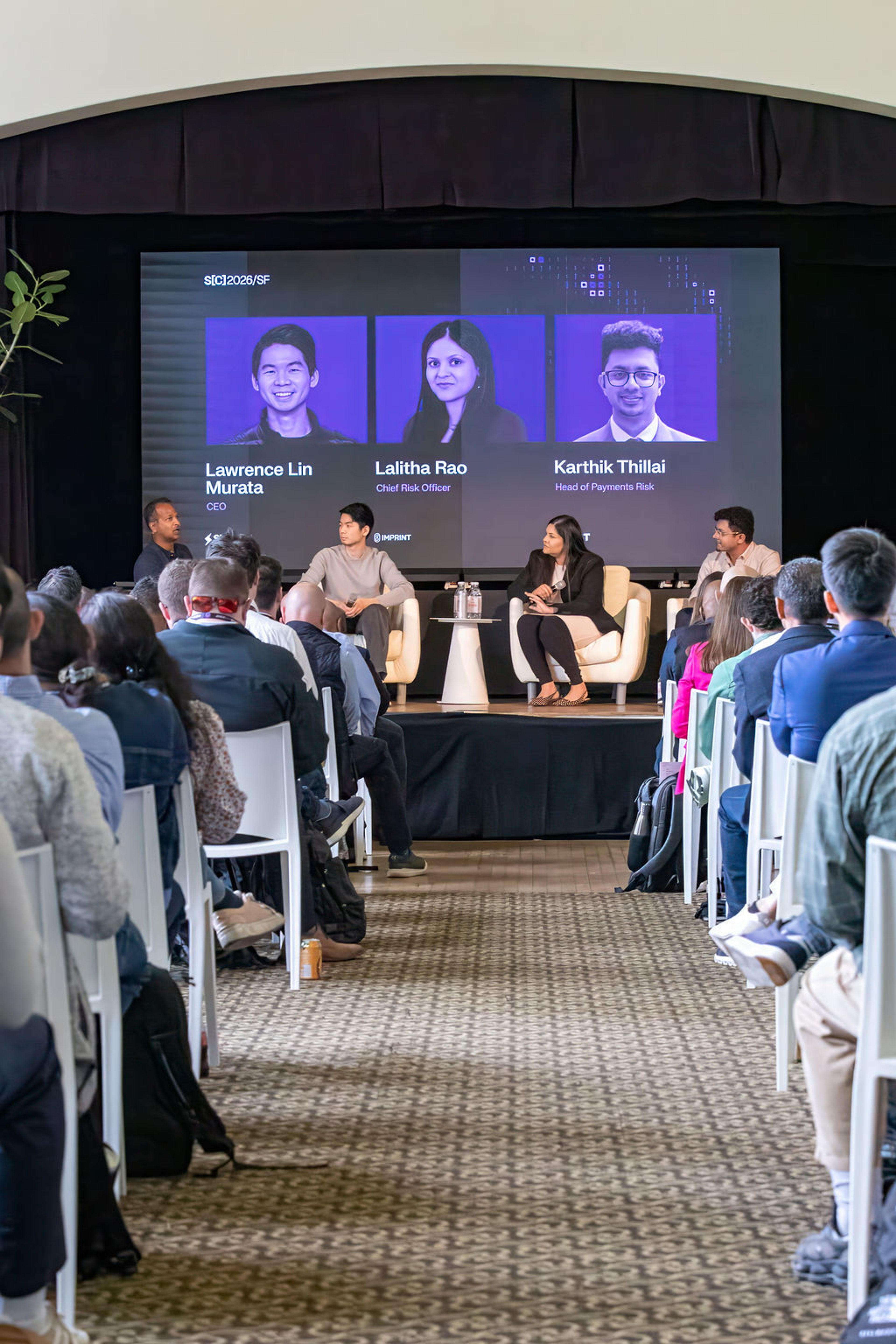 An audience views a panel discussion on a stage with a large screen displaying names and titles of panelists including CEO and Chief Risk Officer.