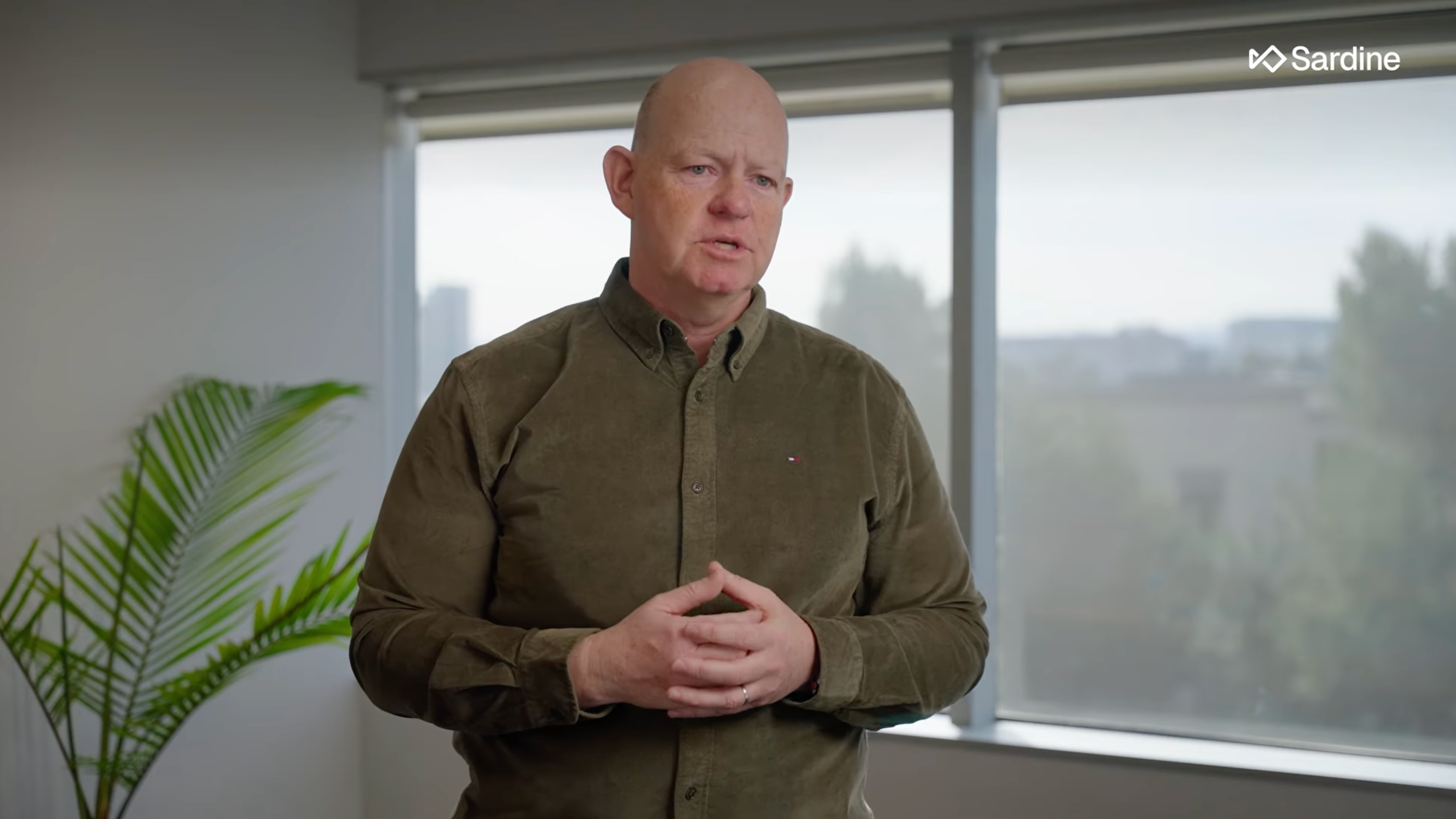 A bald man in a green shirt speaks, hands clasped, in an office with a plant and windows.