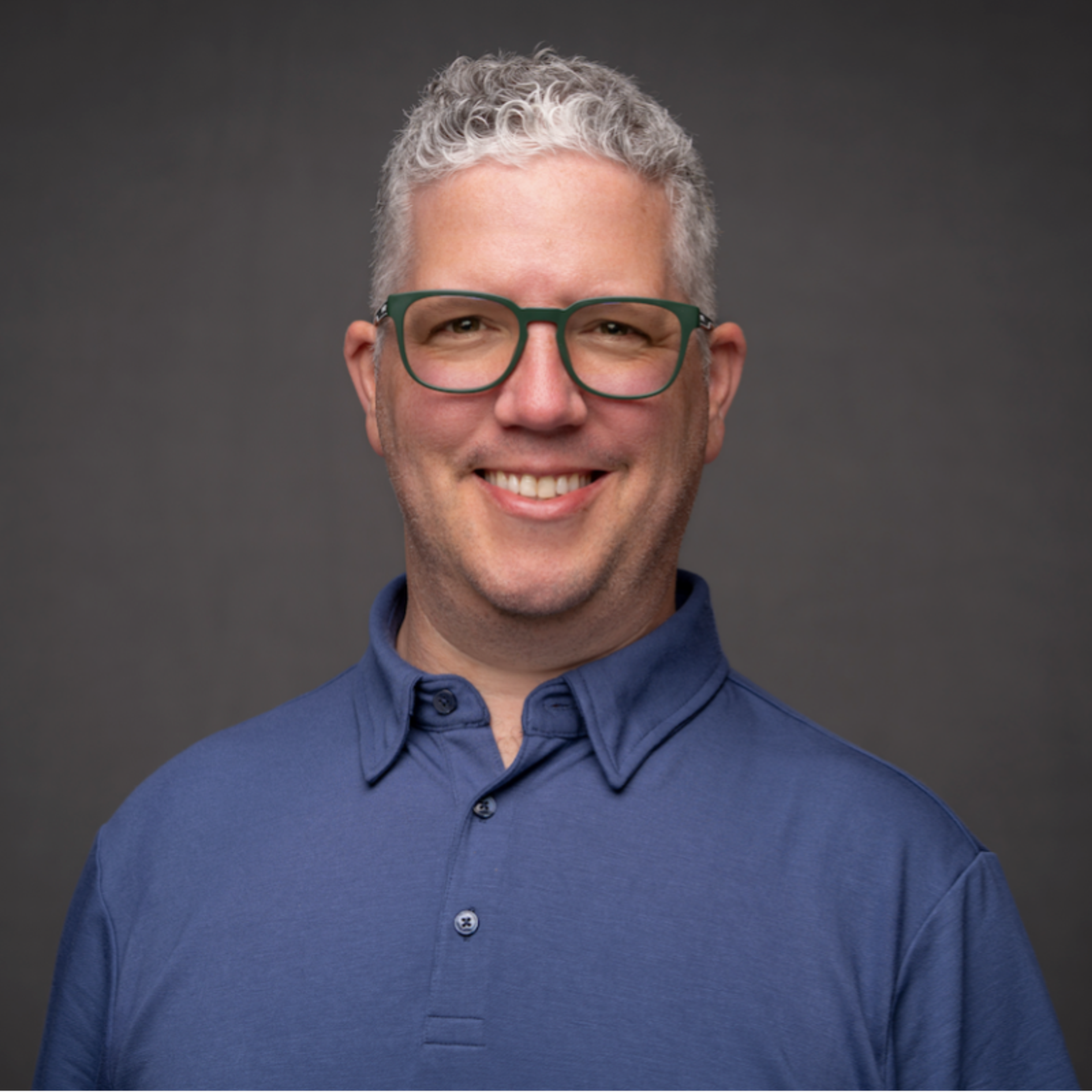 A smiling man with gray hair and green glasses wears a blue polo shirt.
