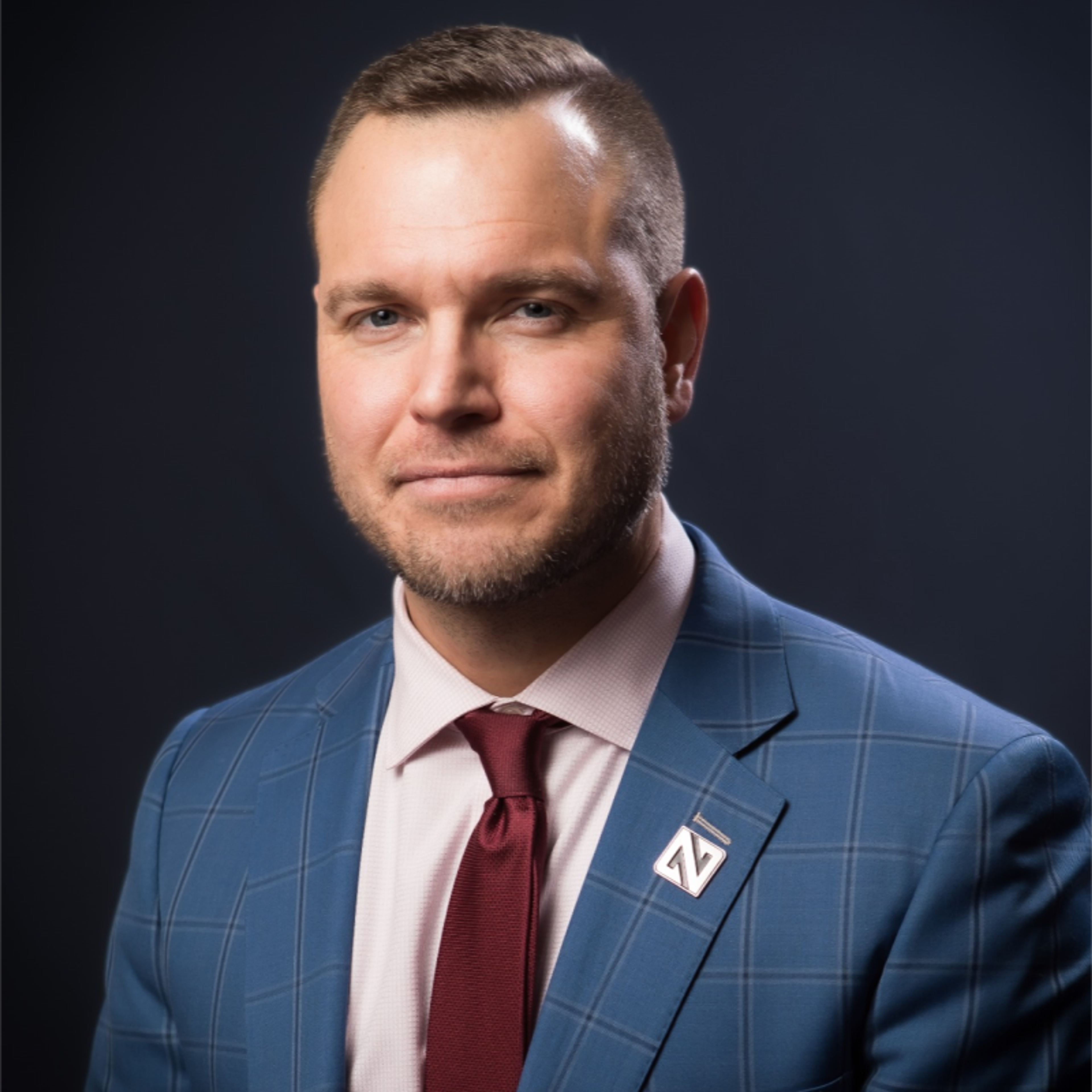 Headshot of a man in a blue plaid suit, pink shirt, and red tie.