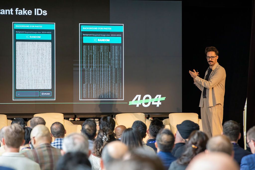 A man presents on stage to an audience, with a screen behind him showing two app interfaces displaying random textured backgrounds, a crossed-out "404," and the partial title "fake IDs."