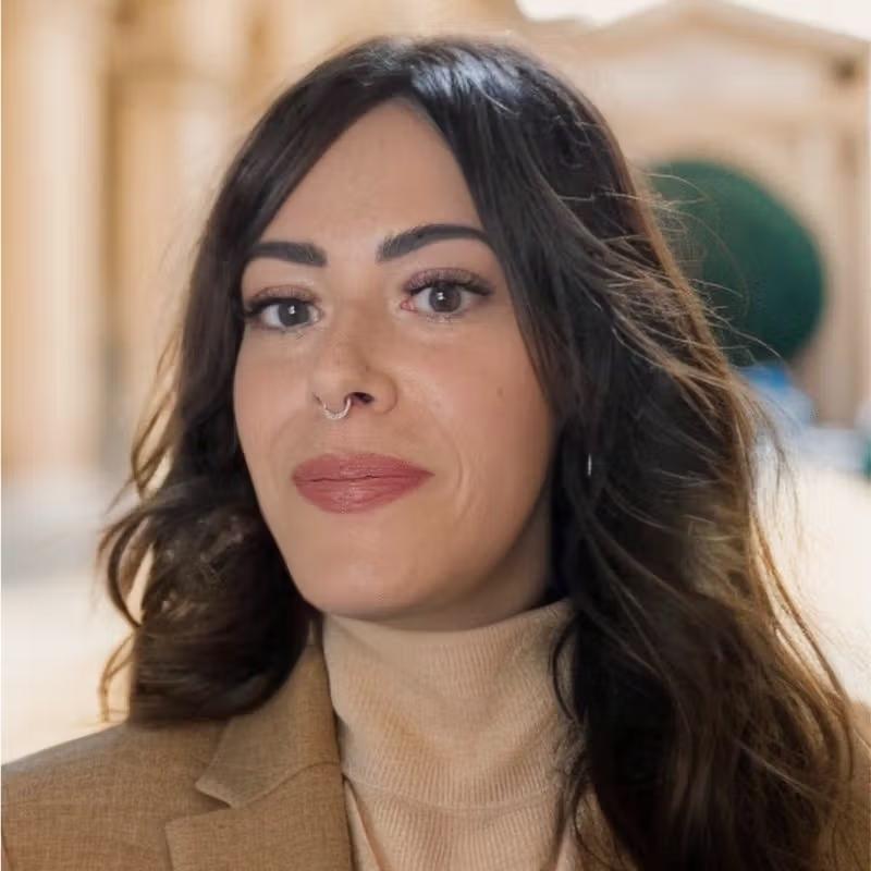 A woman with dark hair, a septum piercing, and a faint smile, wearing a beige turtleneck and brown blazer.