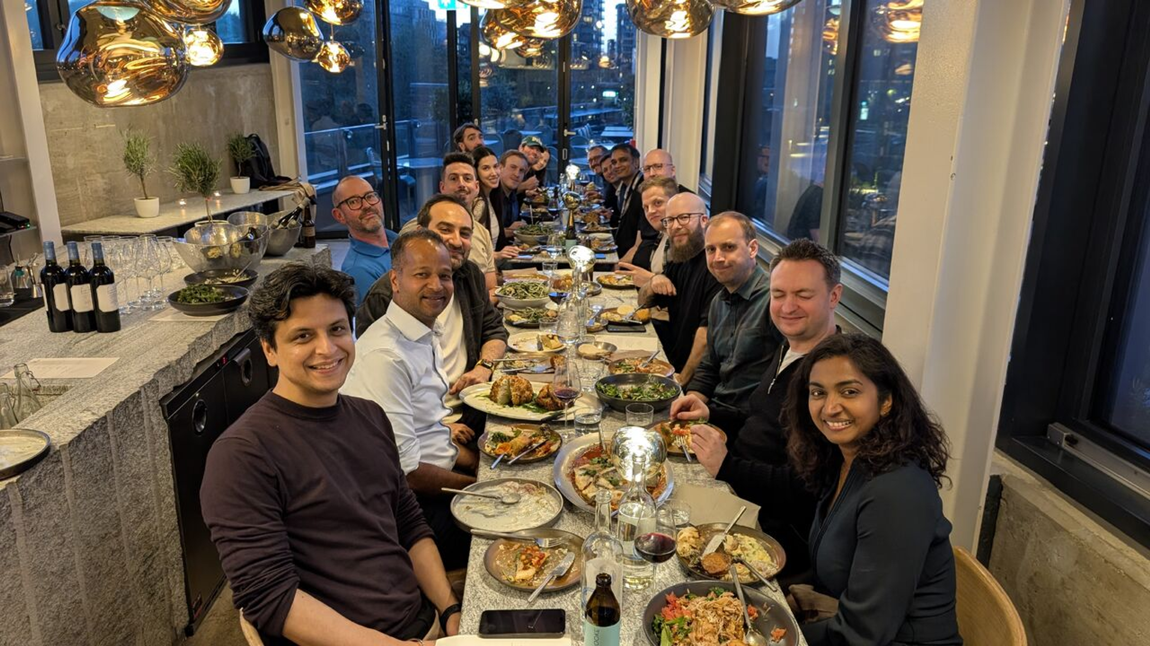 A diverse group of people smiling and dining together at a long restaurant table filled with food.