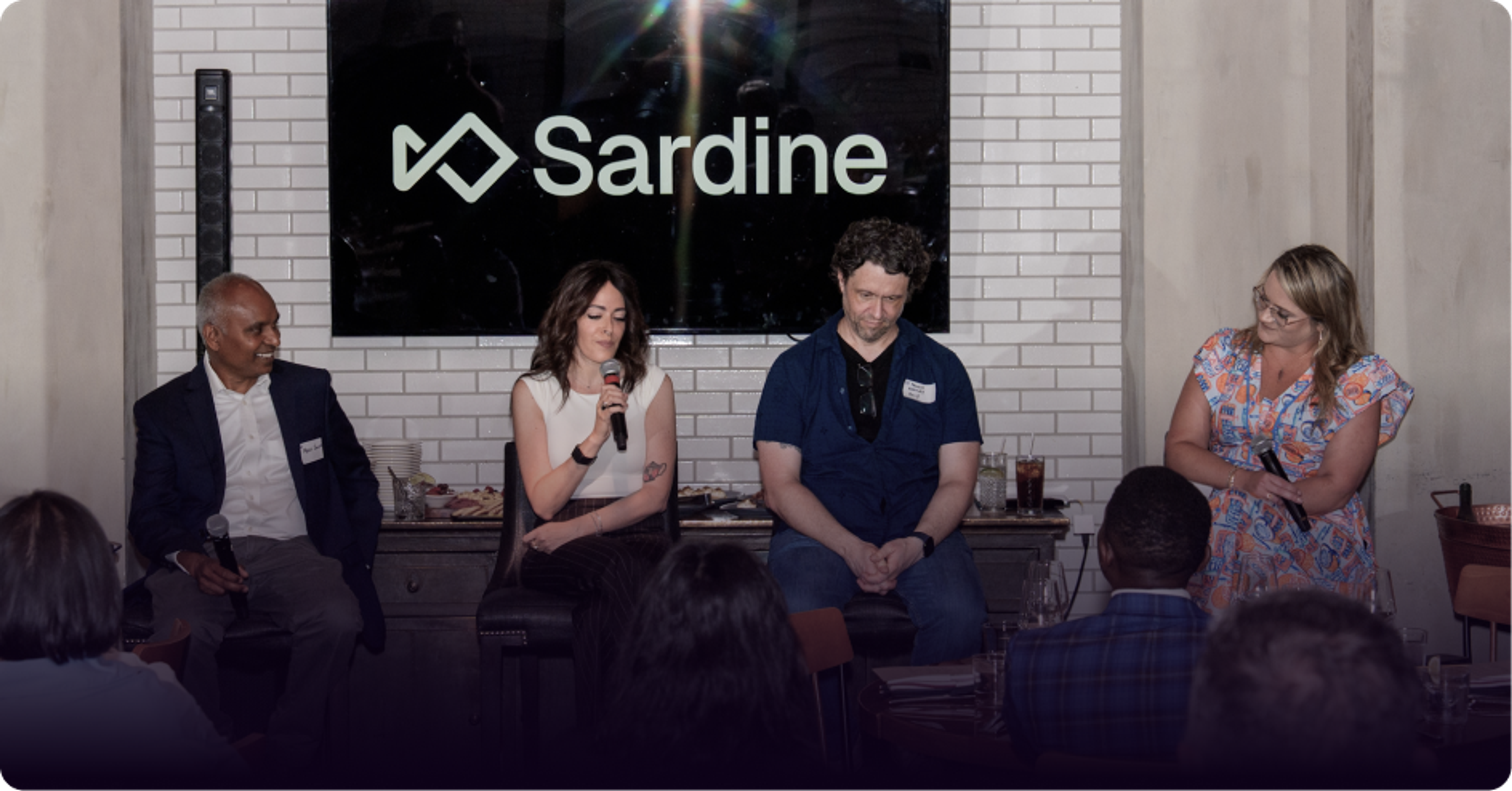 A panel of four speakers, three seated and one standing, in front of a screen displaying the Sardine logo, with an audience.