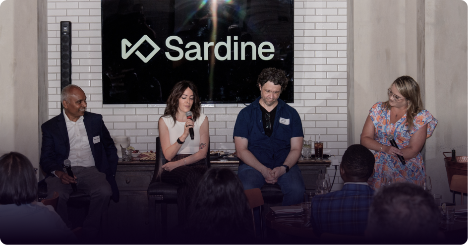 A panel of four speakers, three seated and one standing, in front of a screen displaying the Sardine logo, with an audience.