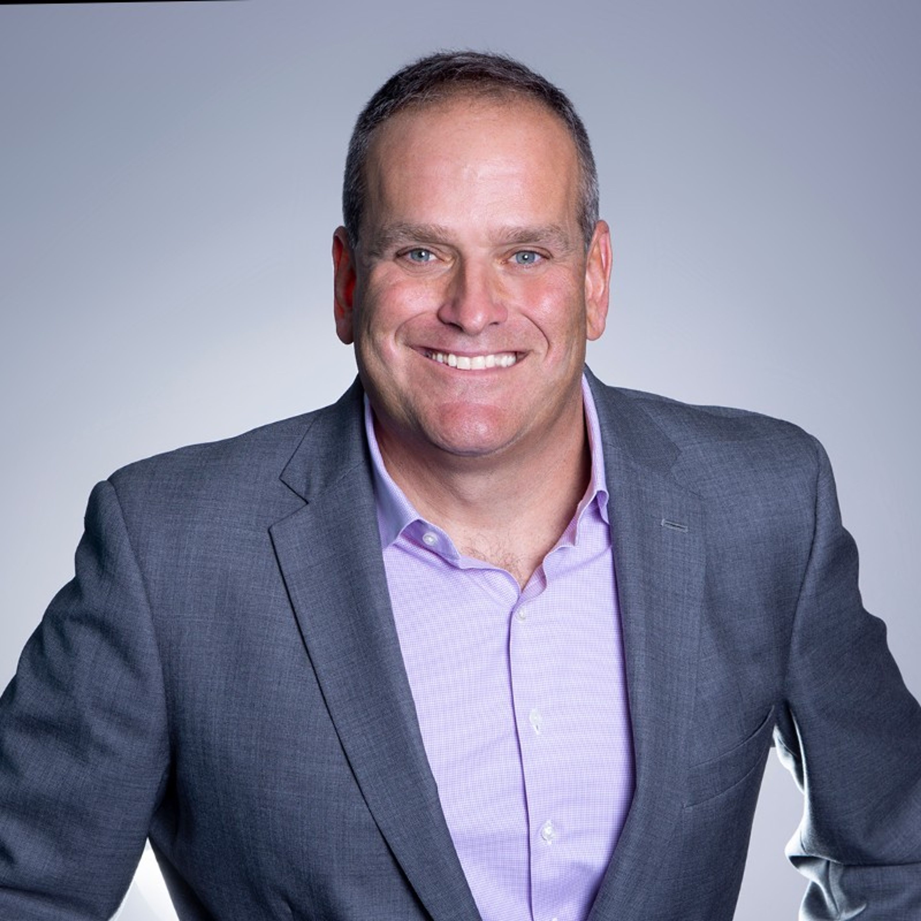 A smiling man in a grey suit jacket and purple shirt.