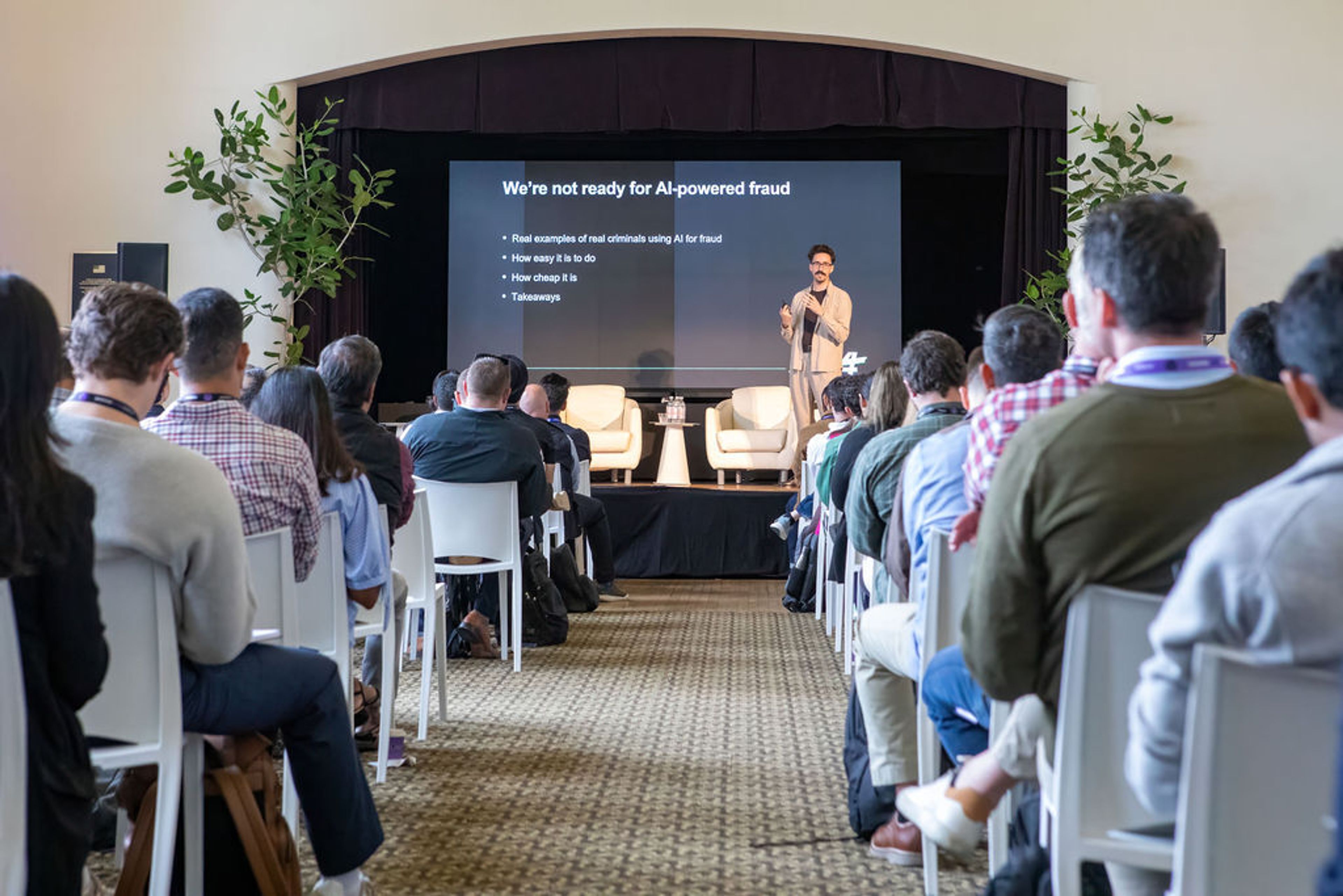 A speaker presents "We're not ready for AI-powered fraud" on a large screen to an audience.