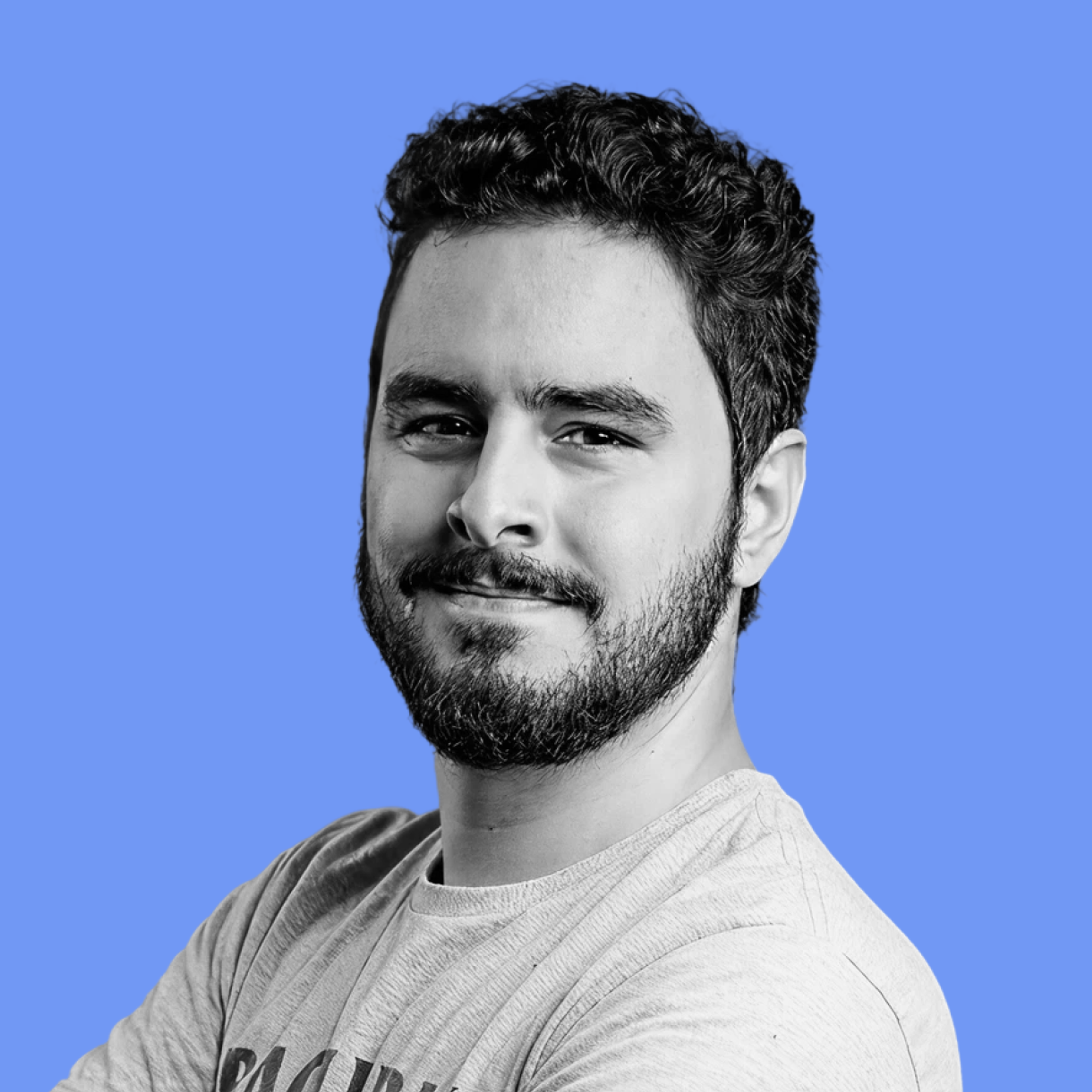 A smiling man with a beard and curly hair, wearing a gray t-shirt, arms crossed, against a blue background.