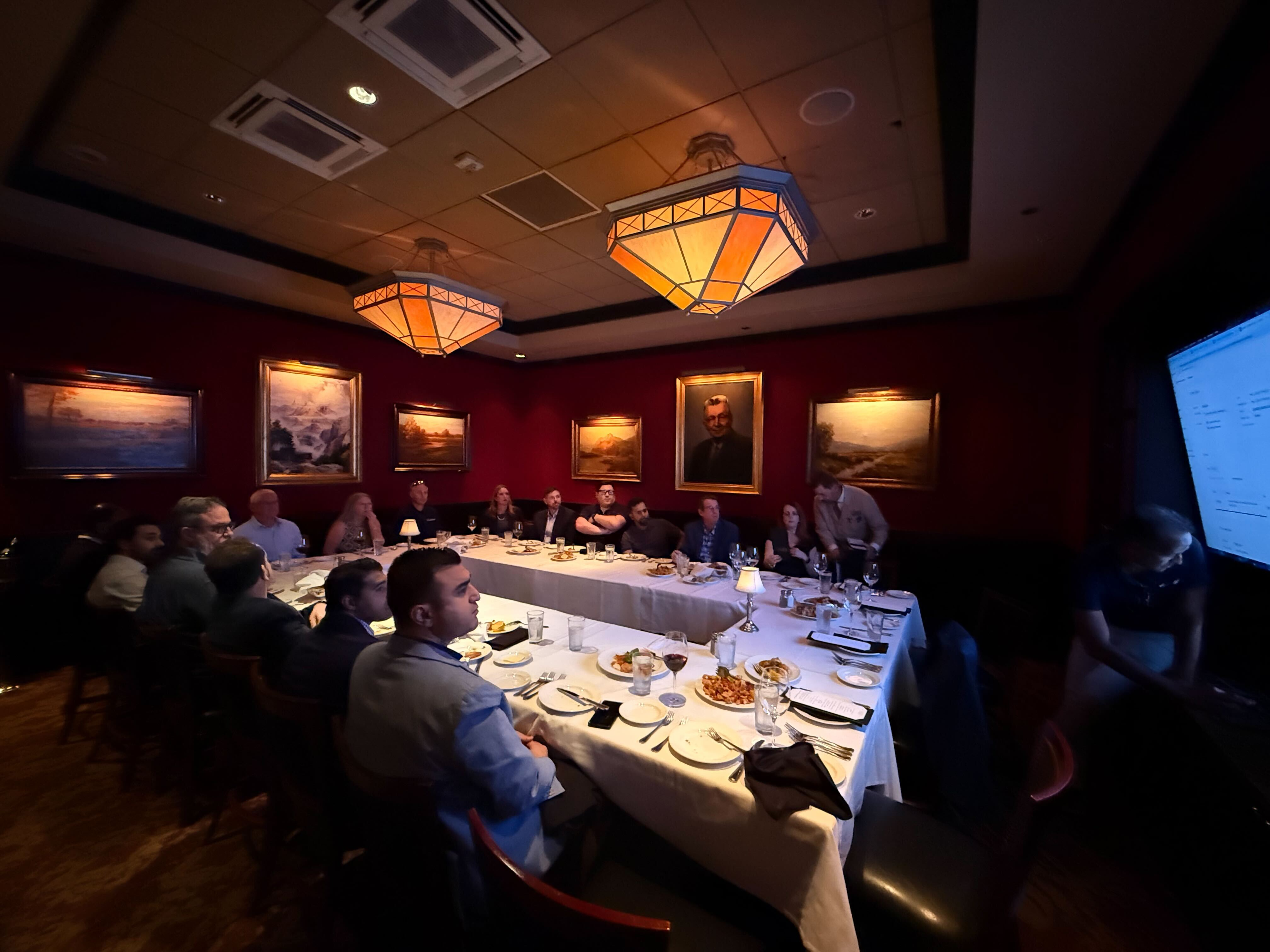 A group of people seated around a long table in a private dining room with red walls, paintings, and a presentation screen.