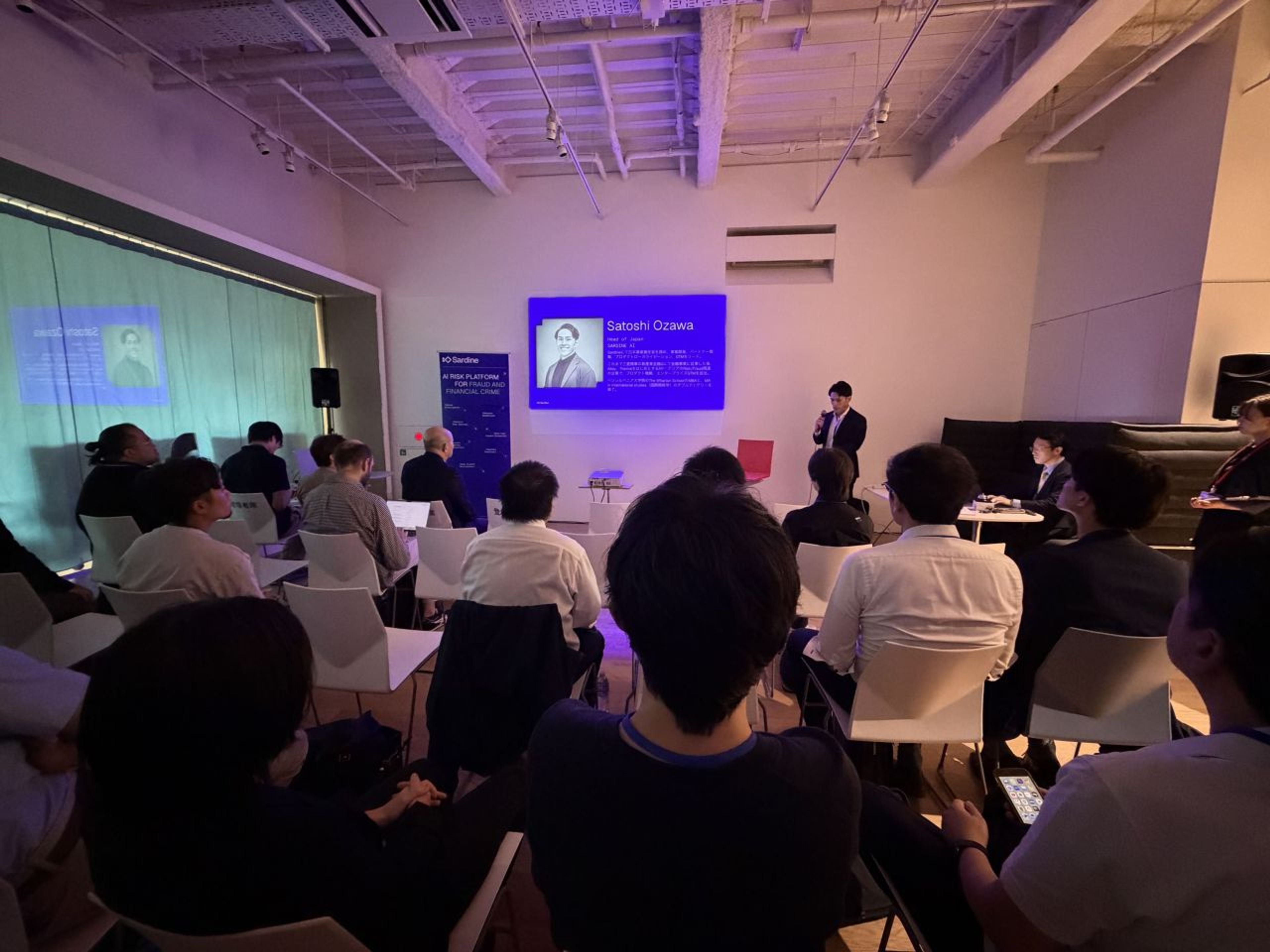 An audience watches a speaker present in front of screens displaying "Satoshi Ozawa."