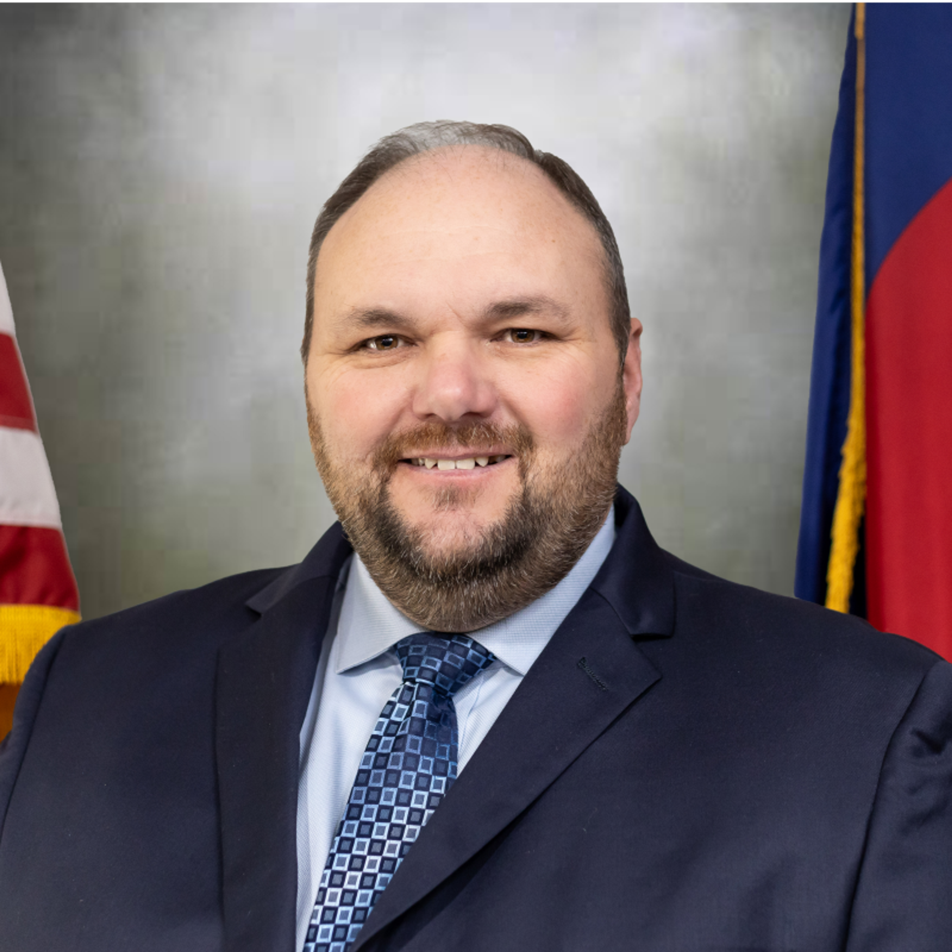 Smiling man with a beard in a suit and tie, with flags behind him.