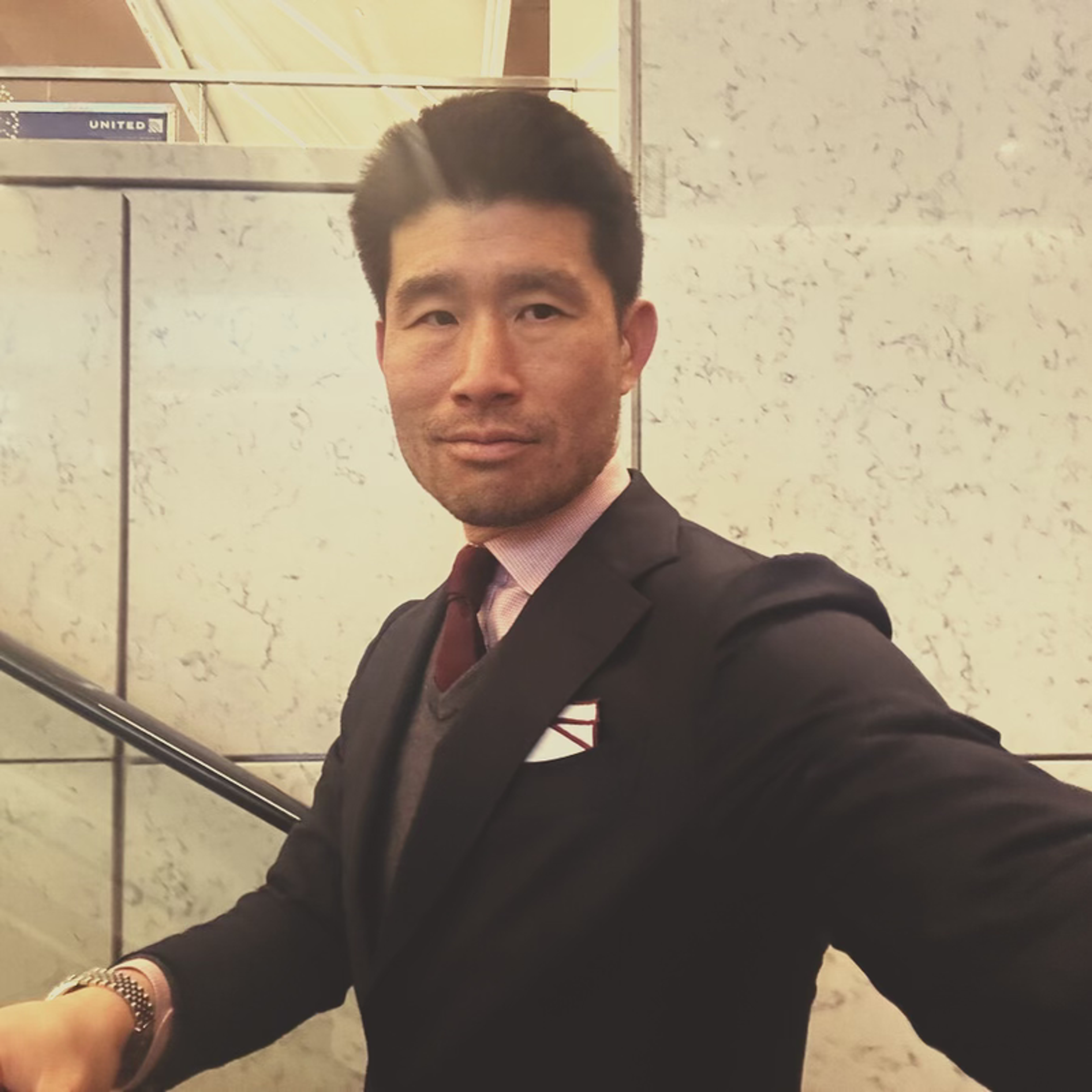 An East Asian man in a navy suit, pink shirt, red tie, and grey sweater on an escalator with a 'UNITED' sign in the background.