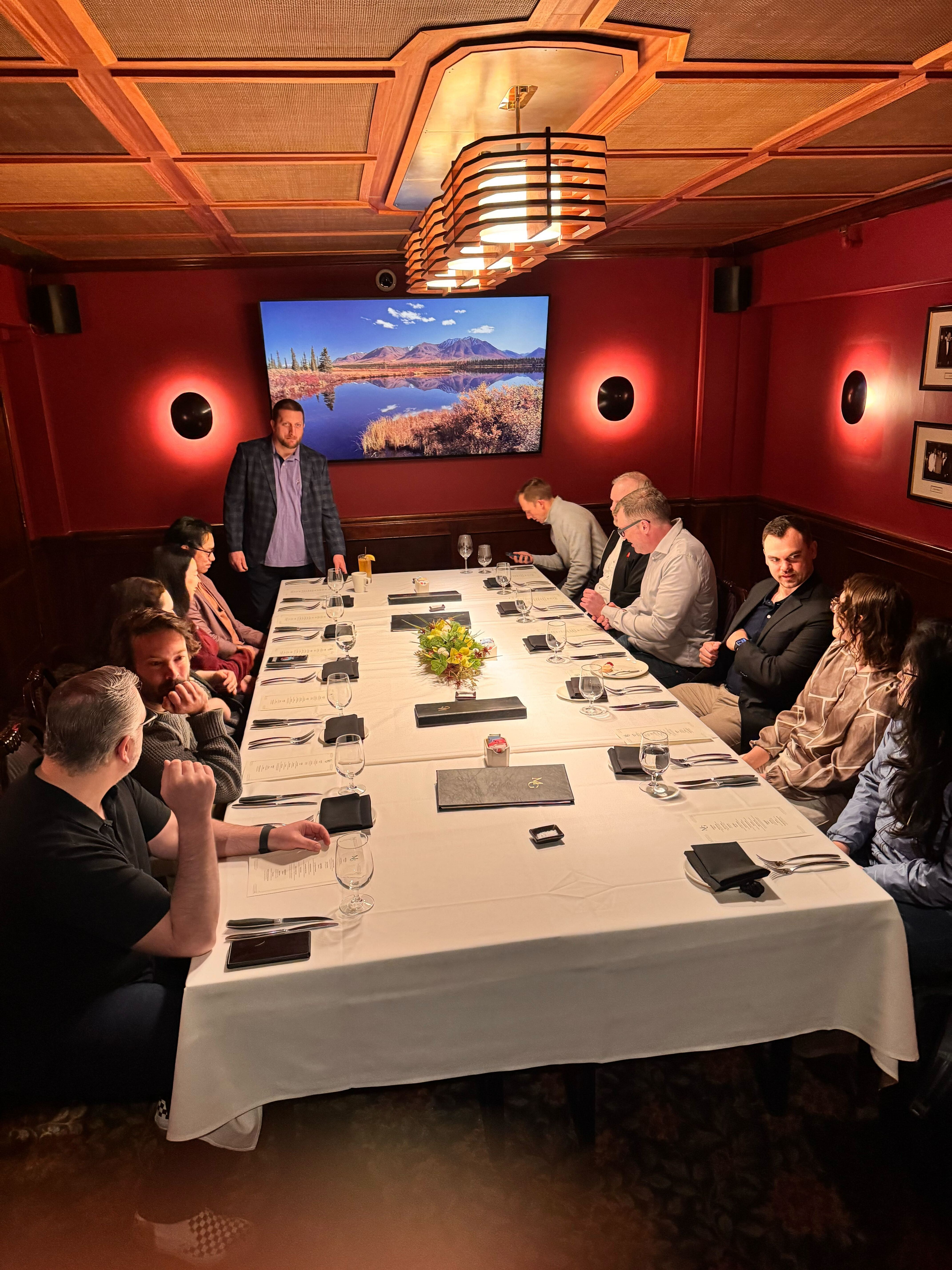 A group of people gathered around a long table in a private dining room, with a man standing in front of a screen displaying a scenic landscape.