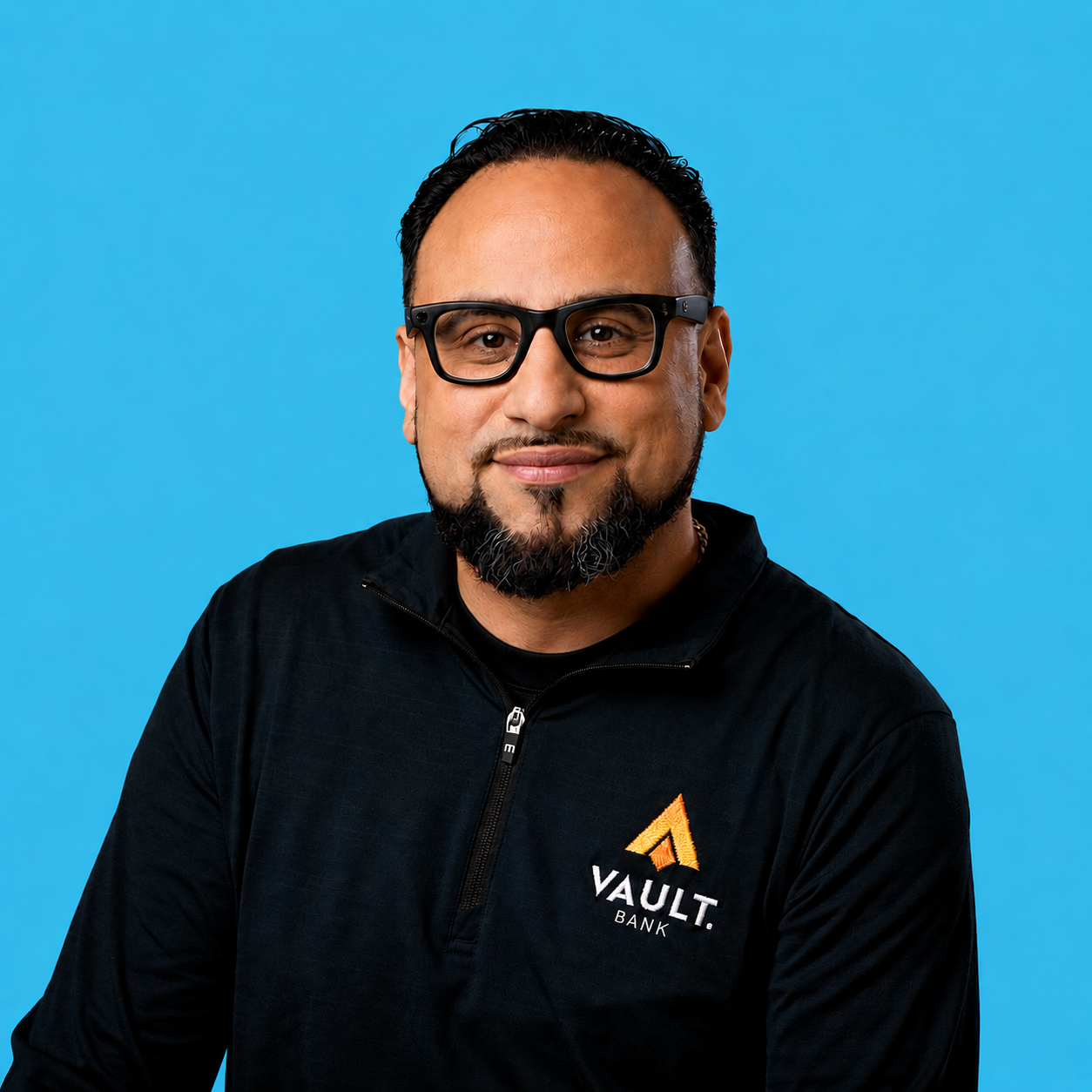 Smiling man with glasses and a beard in a black "VAULT. BANK" shirt on a blue background.