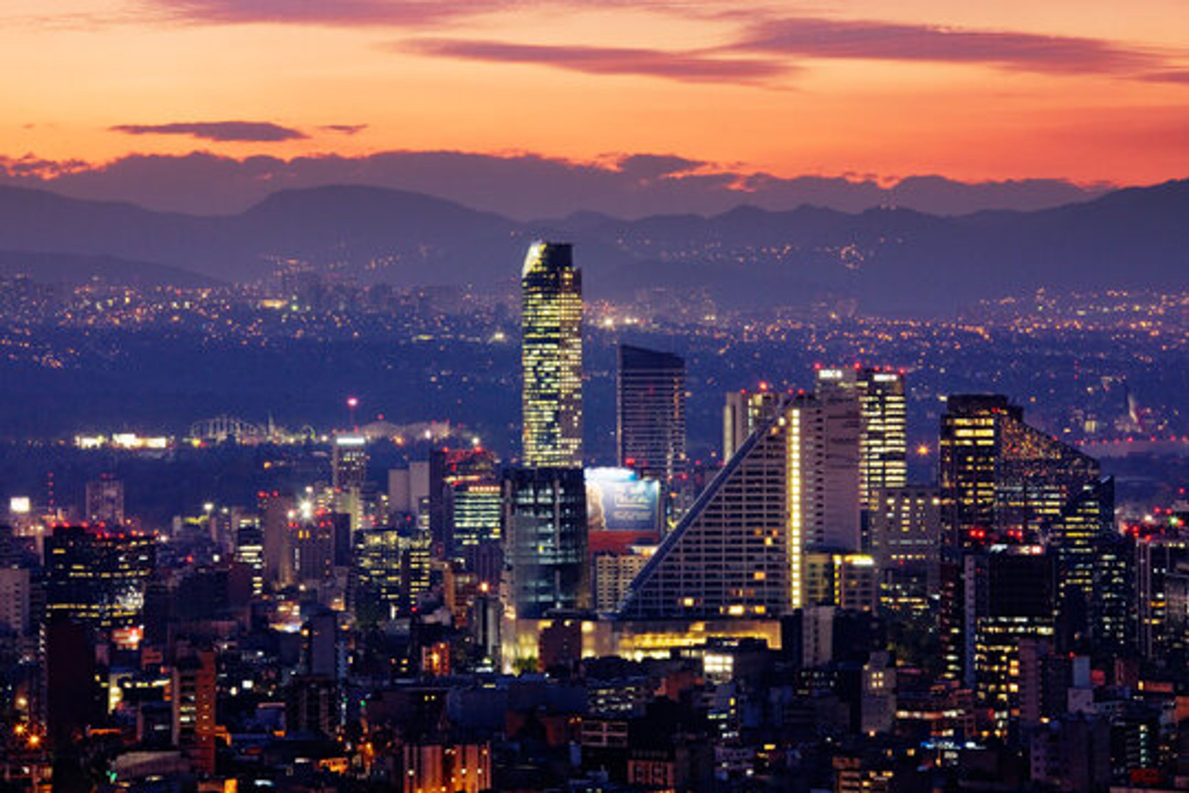 Mexico City skyline at dusk with illuminated skyscrapers and distant mountains.