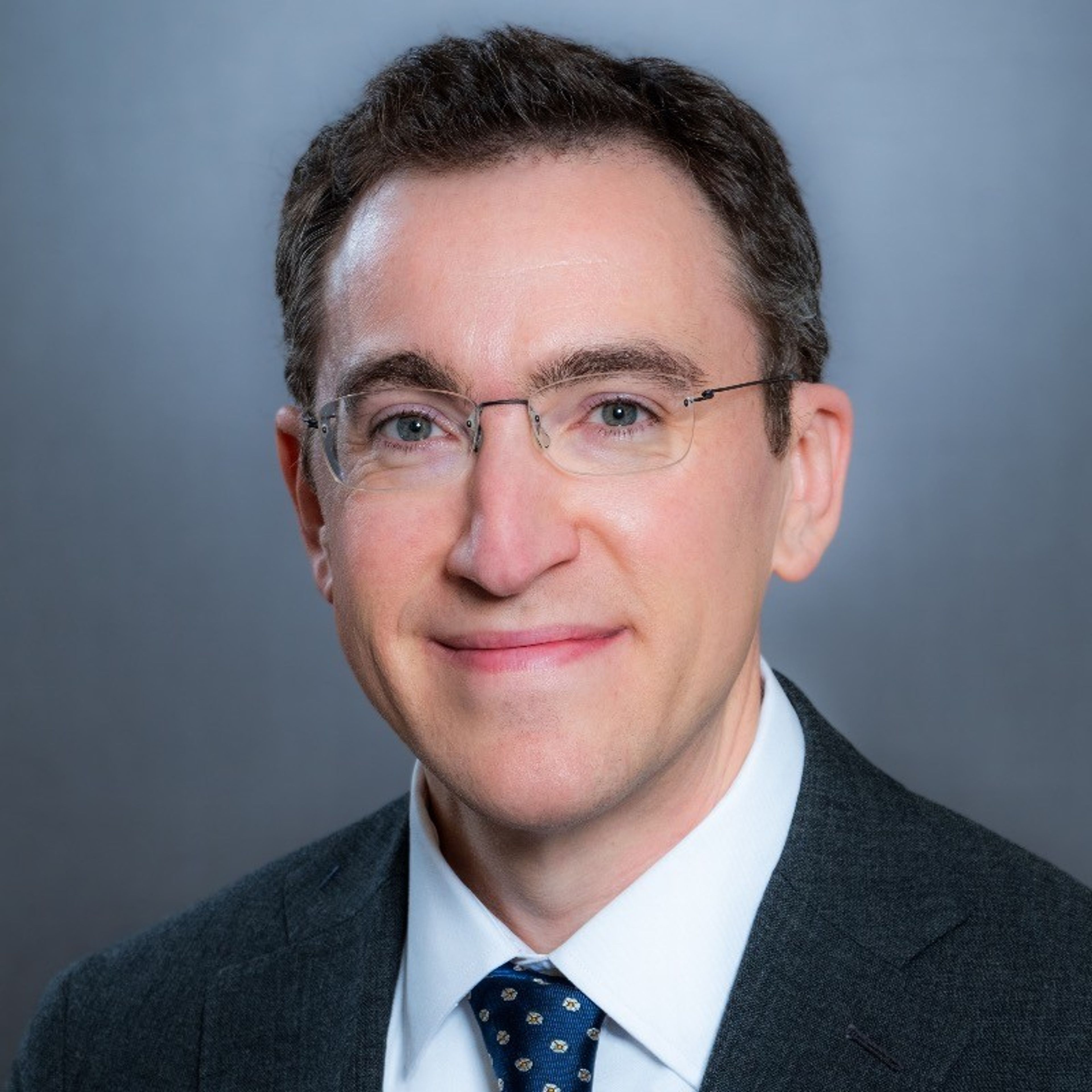 Headshot of a smiling man with glasses in a suit and tie.