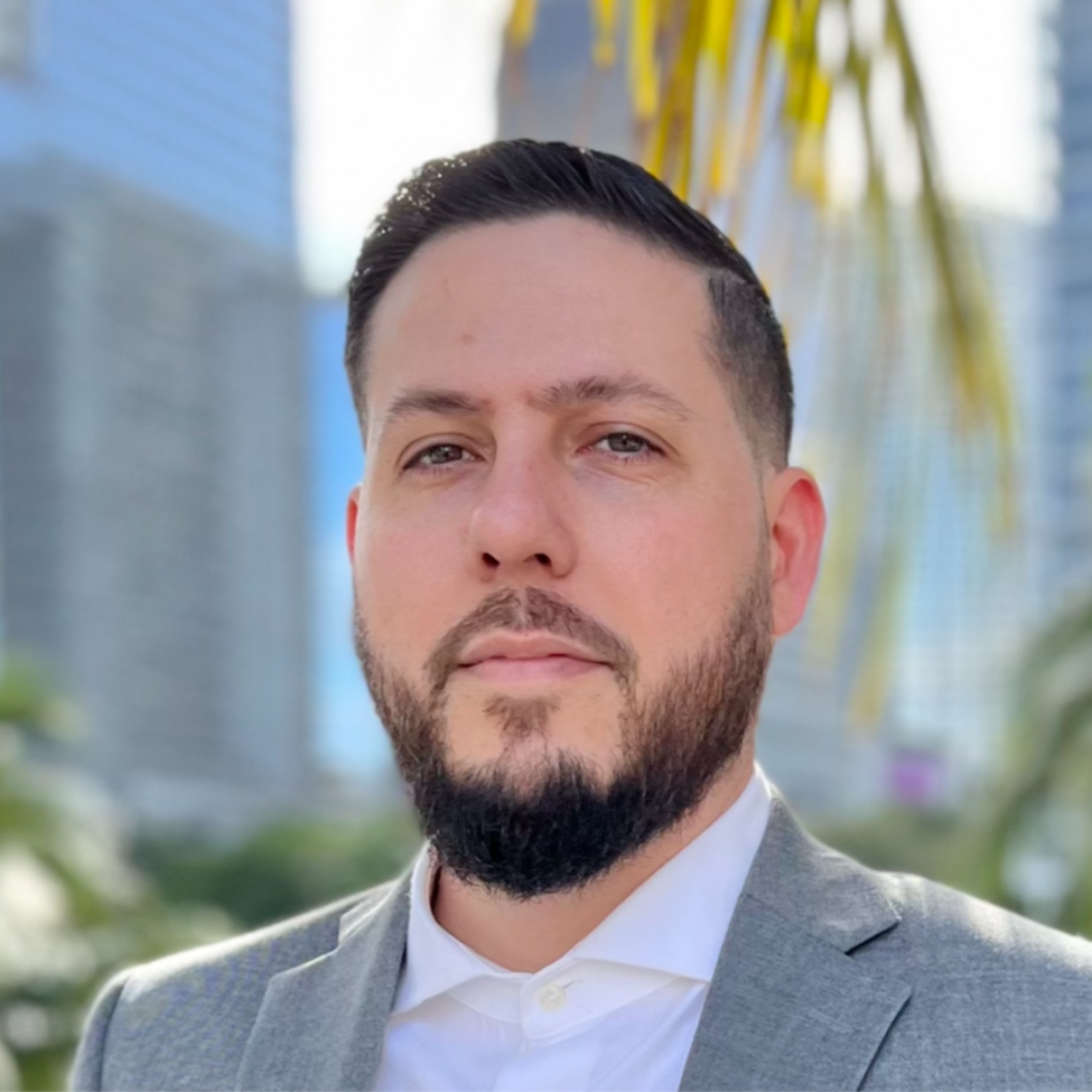Headshot of a man with a dark beard and hair, wearing a grey suit jacket, against an outdoor city background.