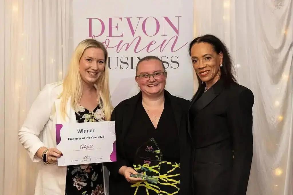 Three women posing for a photo in front of a "DEVON WOMEN IN BUSINESS" backdrop. One woman on the left holds a certificate for "Winner Employer of the Year 2022 Adopstar," and the woman in the middle holds a glass award.