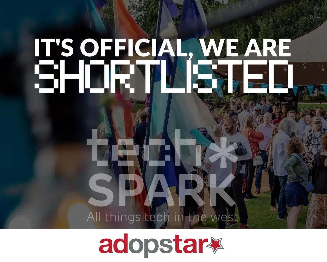 The image features a crowd of people in an outdoor setting, with flags visible in the foreground, against a backdrop of trees and tents. Overlaying this scene in large, bold white text, is "IT'S OFFICIAL, WE ARE SHORTLISTED". Below this, in a faded, pixelated grey font, is "tech* SPARK" and then "All things tech in the west" in a smaller, more legible font. At the very bottom, on a white strip, is the "adopstar" logo in red with a red star.