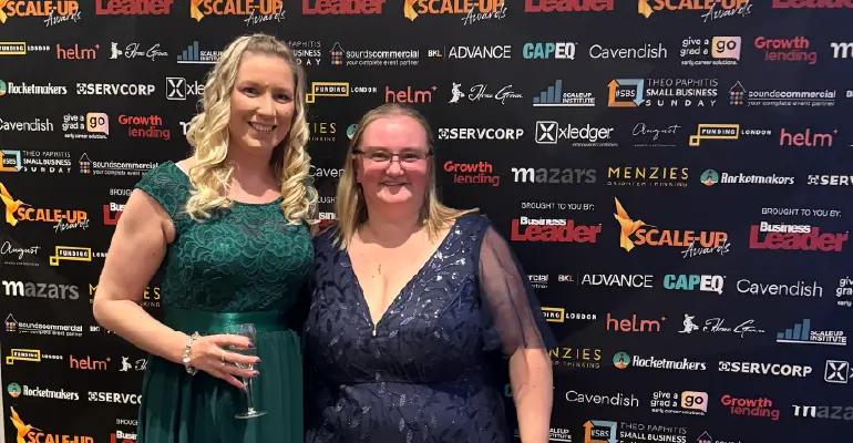 Two women in formal dresses pose in front of a black wall covered with white and gold logos for the "Scale-Up Awards." The woman on the left wears a dark green dress and holds a wine glass, while the woman on the right wears a dark blue dress.