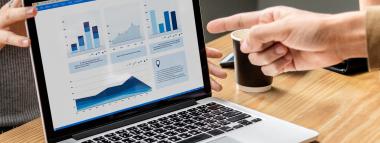  A laptop displays a custom dashboard with various graphs and charts, reflecting analytics and reporting. The background shows a blurred office setting with a person and a window.