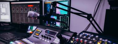 A microphone on a boom arm overlooks a mixing console, surrounded by multiple computer monitors displaying production interfaces in a dimly lit, purple-hued studio. symbolising Radio advertising