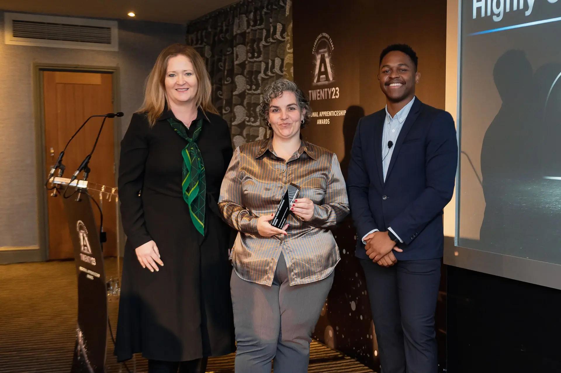 Two women and one man, dressed in smart casual to business attire, pose on a stage. The central woman holds a black award, with a podium to their left and a screen displaying "NATIONAL APPRENTICESHIP AWARDS 23" behind them.