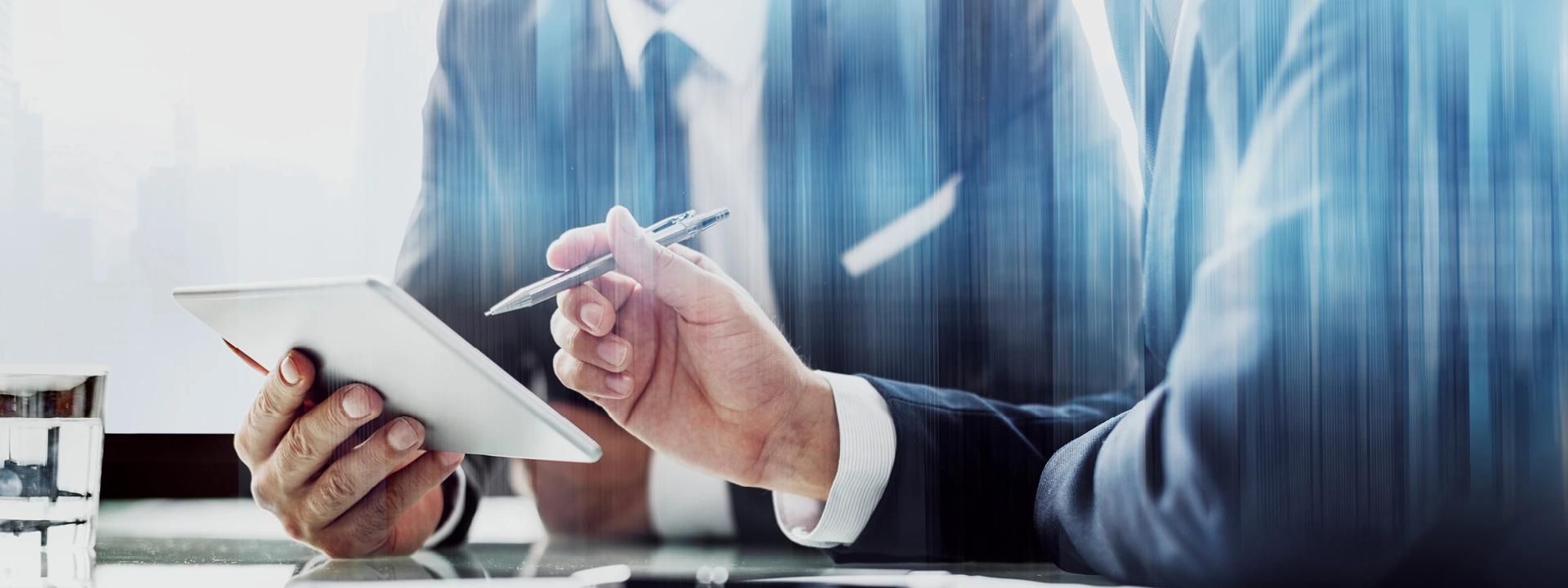 Two professionals in suits are engaged in a discussion, with one holding a tablet and a pen, symbolising a business consultation or meeting.
