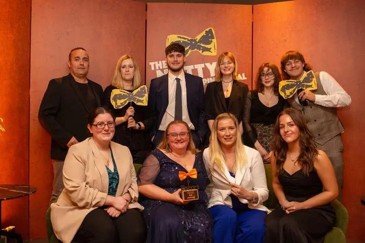short Nine people, dressed formally, are pictured indoors at "The Notty Digital" awards event. Five stand behind four seated individuals; a woman in the front row holds an award featuring an orange bow tie, with two people in the back row holding yellow bow tie cutouts.