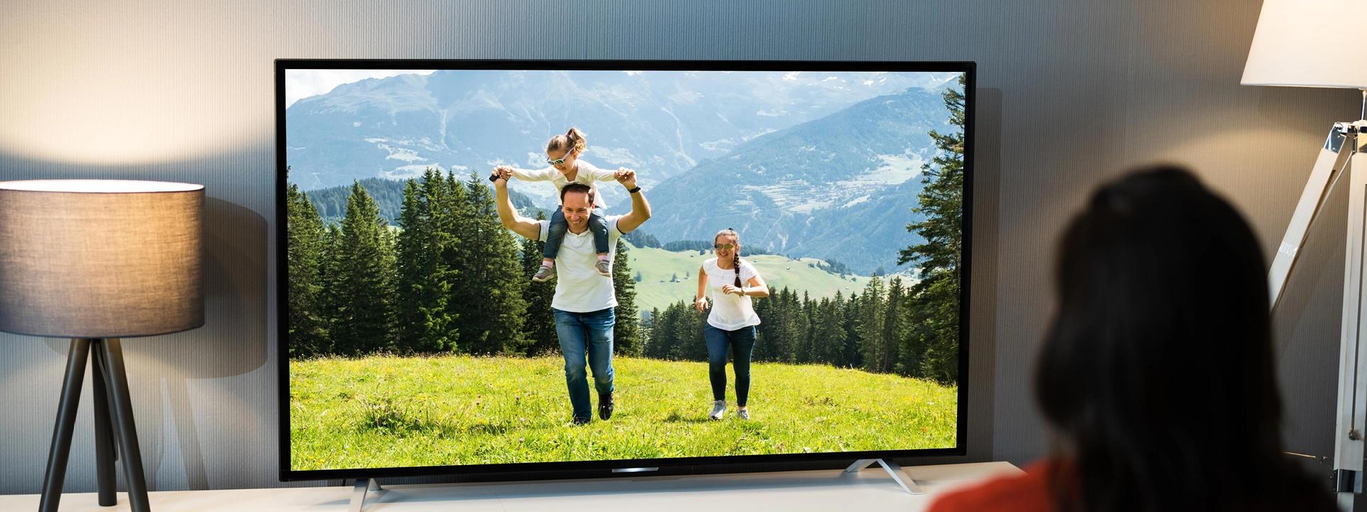  A person views a TV showing a family in a scenic outdoor setting, flanked by two lamps symbolising TV advertising