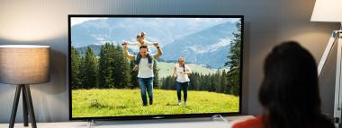  A person views a TV showing a family in a scenic outdoor setting, flanked by two lamps symbolising TV advertising.