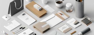  An overhead flat lay showcases a collection of promotional branding items on a white surface, including a grey shopping bag, various brown and white packaging boxes, envelopes, business cards, coffee cups, a jar, pens, pencils in a holder, and small tech gadgets, all uniformly styled.