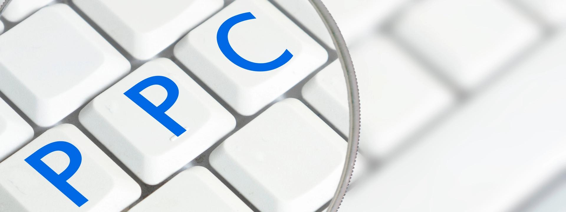 A magnifying glass is positioned over a white computer keyboard, clearly showing the blue letters 'PPC' on three individual keys, symbolising a focus on Pay-Per-Click advertising for B2B businesses.