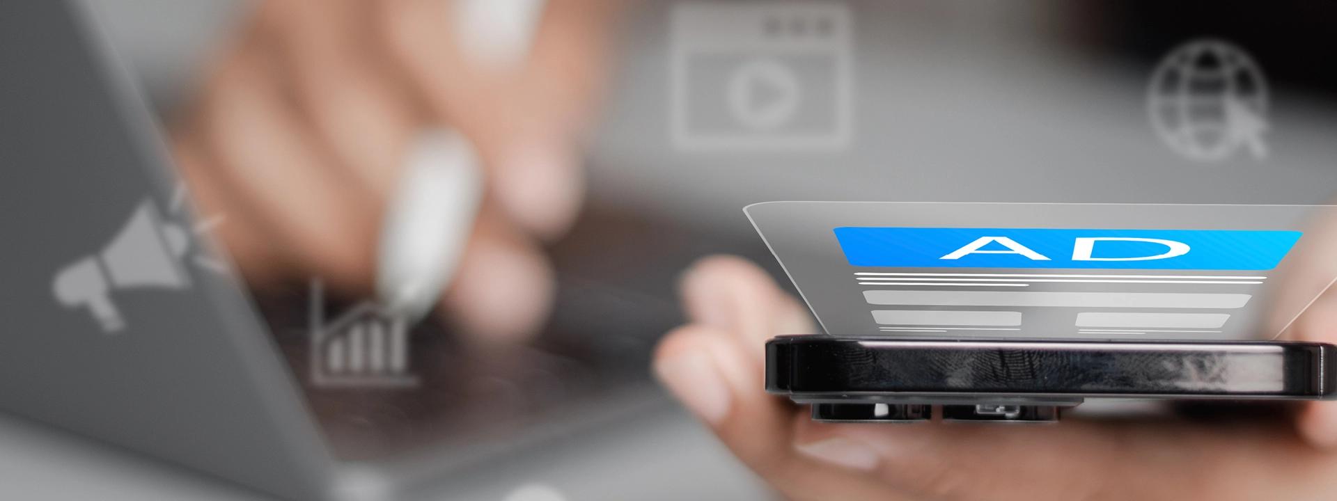 A close-up of a hand holding a smartphone displaying an "AD" label on its screen, with blurred digital marketing icons like a megaphone and a graph in the background, representing paid social media advertising.