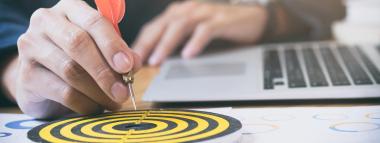 A person's hand is holding a red-feathered dart, poised to hit a small yellow and black target on a desk next to a laptop, symbolising the precision and targeting involved in retargeting and remarketing advertising.
