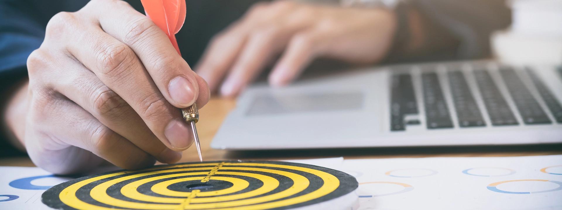 A person's hand is holding a red-feathered dart, poised to hit a small yellow and black target on a desk next to a laptop, symbolising the precision and targeting involved in retargeting and remarketing advertising.