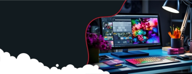  A desktop computer monitor displays vibrant graphic design software with colorful images and a grid layout. A brightly lit desk lamp, keyboard with colorful backlighting, mouse, and a purple floral arrangement are also visible on a blue desk. The left side of the image is a dark background with a red wavy line and white cloud shapes.