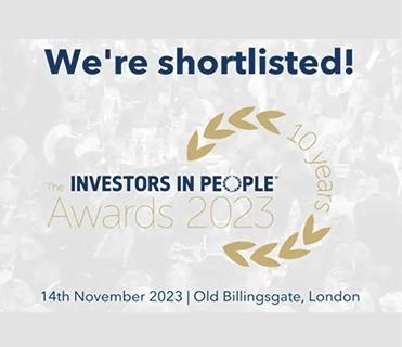 The image is a graphic on a light background, announcing "We're shortlisted!" in large dark blue text at the top. Below, a faded crowd image serves as a subtle background for the "The INVESTORS IN PEOPLE® Awards 2023" logo in grey, partially encircled by a golden wreath-like design with "10 Years" written alongside. At the bottom, the event details "14th November 2023 | Old Billingsgate, London" are displayed.