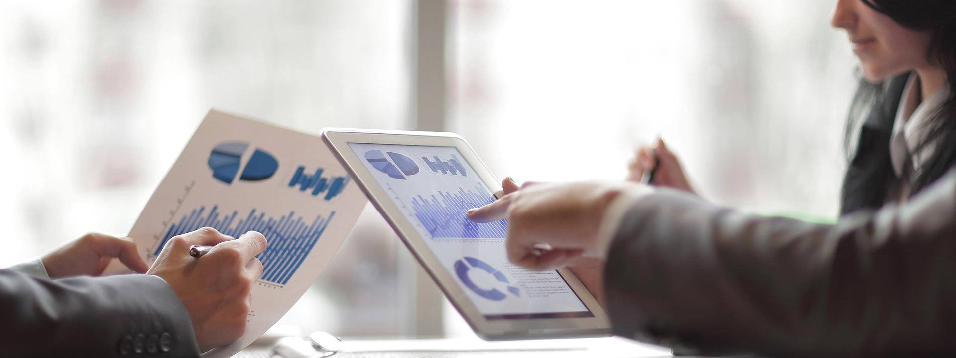 Two people reviewing competitor analysis data on a tablet and printed charts, with one person pointing at the tablet screen.