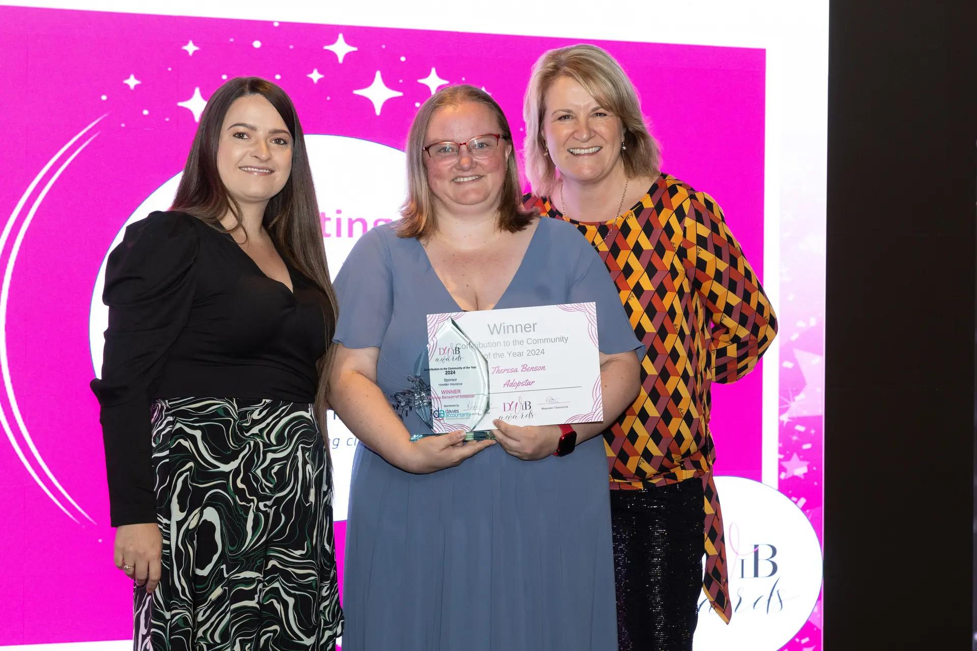 Three women are smiling and standing in front of a bright pink backdrop with event branding. The woman in the center is holding a glass award plaque and a certificate that reads 'Winner Devon Women in Business Contribution to the Community of the Year 2024' for 'Theresa Benson Adopstar'.