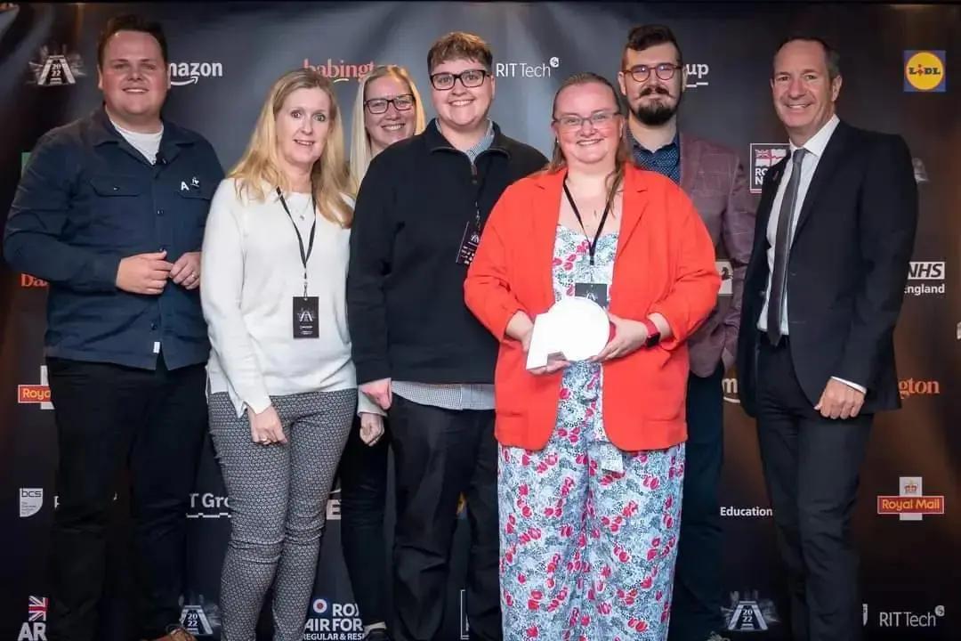 A group of seven people, including a woman in a red blazer holding a white award where Adopstar won Regional SME Employer of the Year smiling and posing in front of a sponsored backdrop at an event.