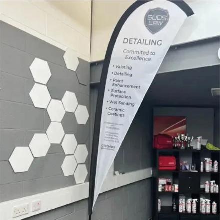  A tall, teardrop-shaped "SUDS LAW DETAILING" feather banner stands indoors, listing services like valeting and ceramic coatings. Behind it, a grey wall features a hexagonal pattern, and shelves on the right display car detailing products.