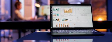  A laptop displaying various graphs and charts on its screen, indicative of data analytics and reporting, sits on a reflective surface. The background is blurred, showing an office environment with a person working and a window with a sunset glow.