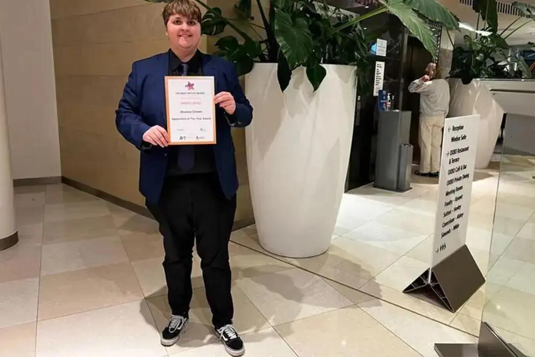 shorten A person with short dark hair, dressed in a blue suit and black attire, stands on a tiled floor, holding up an "Apprentice of the Year Award" certificate and smiling at the camera. A large white planter with green foliage is to their right, and a directional sign is visible further back.