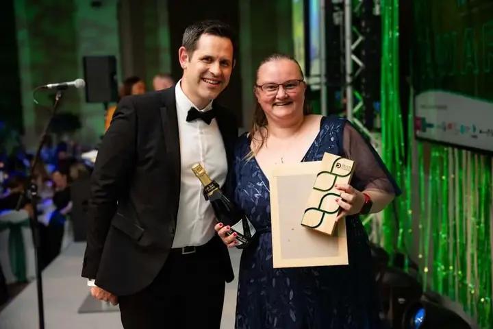 A man in a tuxedo holding a bottle of champagne and Theresa in a blue dress holding a Devon & Plymouth Chamber Awards 2024 - The Rapid Riser, both smiling at the camera