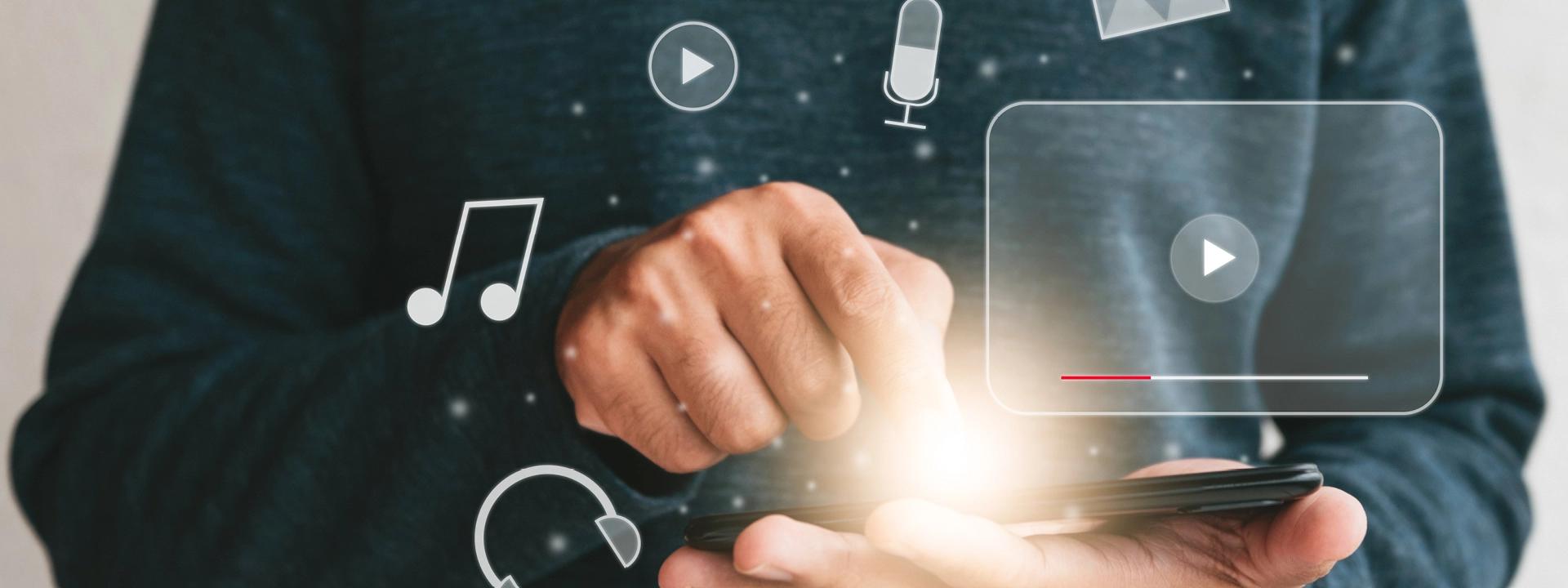 A person holding a smartphone, with glowing light emanating from the screen, surrounded by holographic icons representing various multimedia elements like play buttons, musical notes, a microphone, and headphones, symbolising multimedia design and interaction.