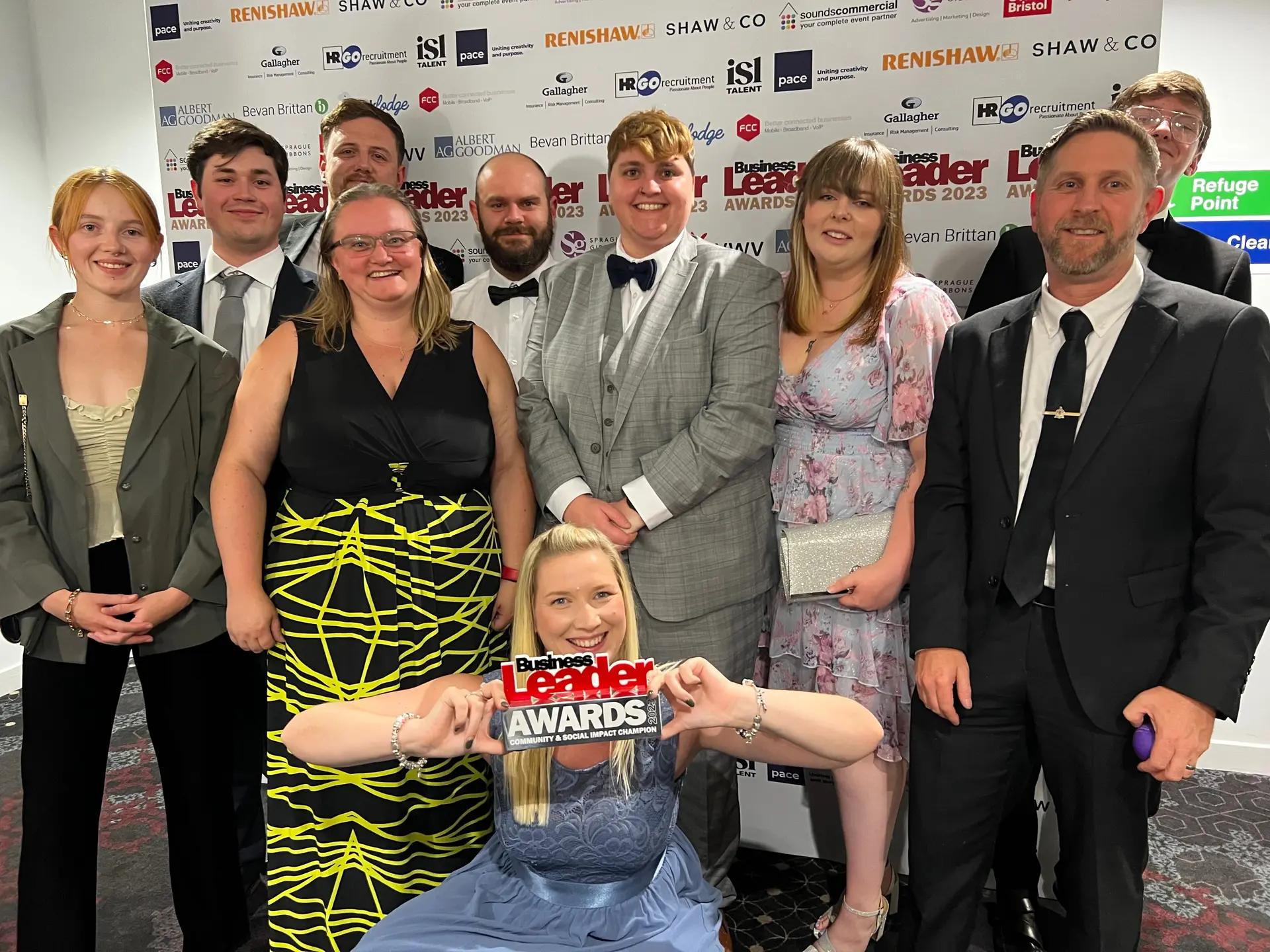 A group of ten formally dressed individuals, likely at an awards ceremony, pose for a photo. A woman seated on the floor holds a "Business Leader Awards" sign, while nine others stand behind her, all smiling amidst event signage symbolising Adopstar as a finalist in the Business Leader Awards