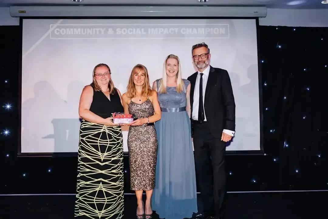 Four people standing on a stage, three women and one man, posing with an award in front of a screen displaying the Business Leader 2023 "COMMUNITY & SOCIAL IMPACT CHAMPION won by Adopstar