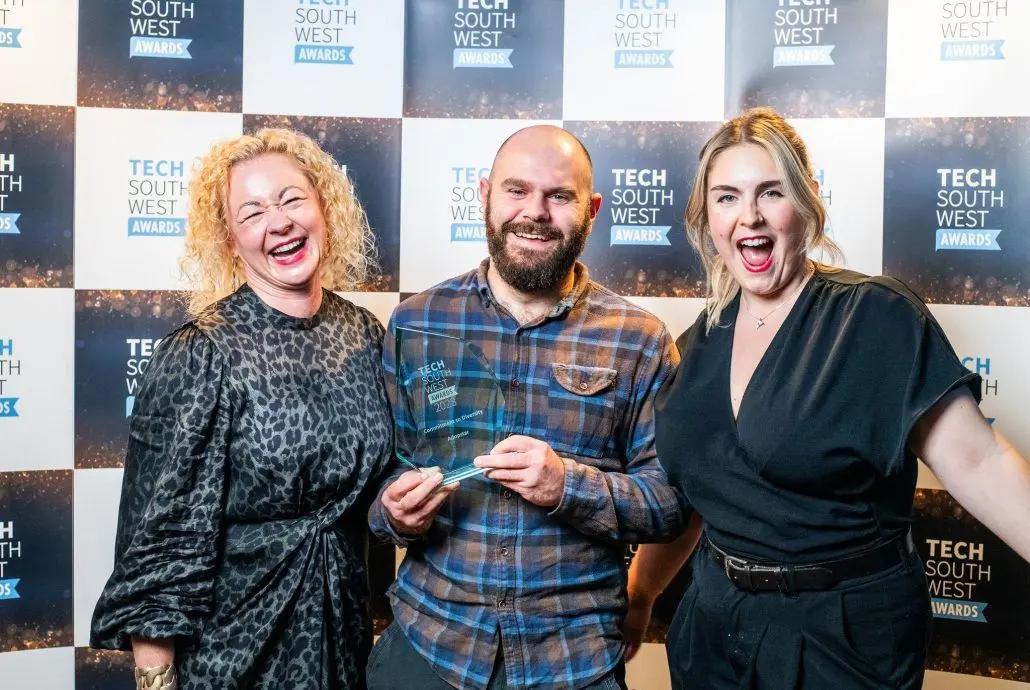 A smiling man in a plaid shirt holds a clear award while standing between two smiling women. They are in front of a backdrop with "TECH SOUTH WEST AWARDS" repeated. Showing the winners of th Winner of the Commitment to Diversity Award at the Tech South West Awards 2023